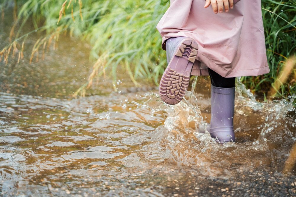 Little girl in pink waterproof raincoat, purple rubber boots funny jumps through puddles on street road in rainy day weather. Spring, autumn. Children's fun after rain. Outdoors recreation, activity