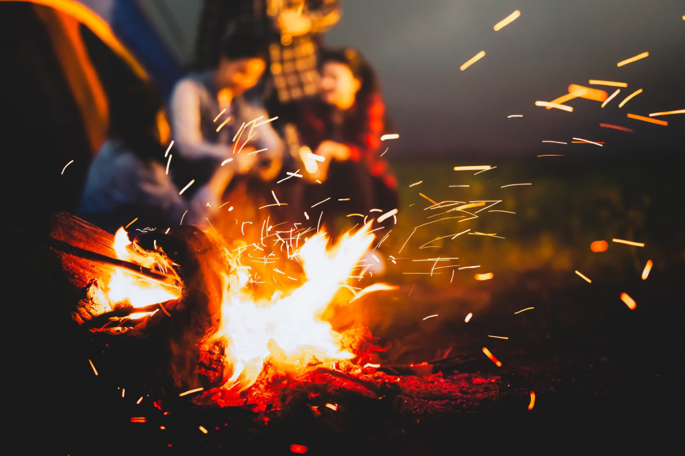 Sparking bonfire with tourist people sit around bright bonfire near camping tent in forest in summer night background. Group of student at outdoor fire fuel. Travel  activity and long vacation weekend