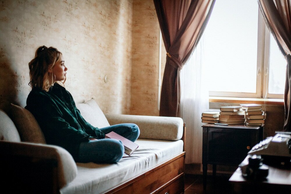 Young sad woman holding book looking through window while sitting on sofa in living room at home